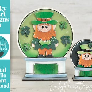 Könnte beinhalten: Zwei Holzdekorationen zum St. Patrick's Day. Eine ist ein großes Schneekugel-Design mit einem Leprechaun, Kleeblättern und dem Text "Pinch Me". Die andere ist eine kleinere Version mit dem Text "Lucky". Das Bild enthält auch den Text "Lucky Heart Designs Co" und "Digital cut file instant download".