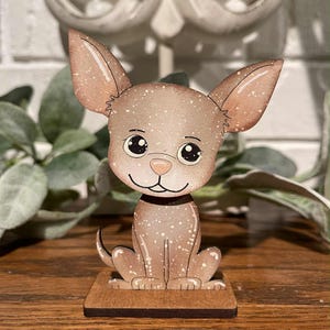 May include: A wooden Chihuahua figurine with a light brown speckled finish. The dog has large ears, a black collar, and is sitting on a small, rectangular wooden base. The figurine has a cartoonish design with large, expressive eyes.