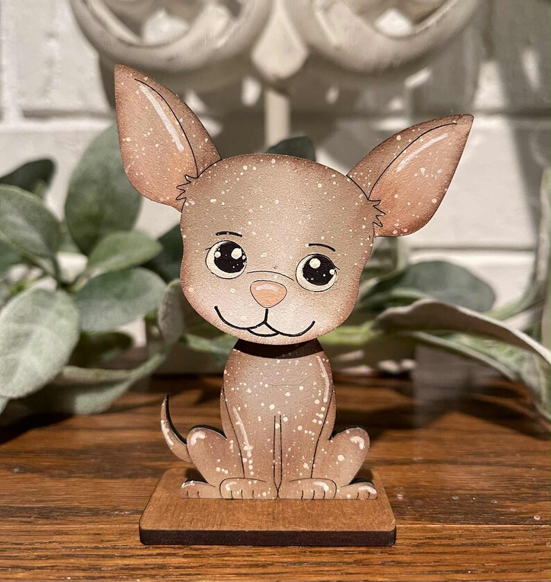 May include: A wooden Chihuahua figurine with a light brown speckled finish. The dog has large ears, a black collar, and is sitting on a small, rectangular wooden base. The figurine has a cartoonish design with large, expressive eyes.