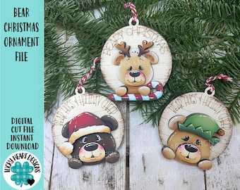 May include: Three wooden Christmas ornaments featuring bear designs. One bear wears a Santa hat, another a green elf hat, and the third has reindeer antlers. Each ornament has a red and white string for hanging and the text "Bear Hugs & Holiday Cheer".
