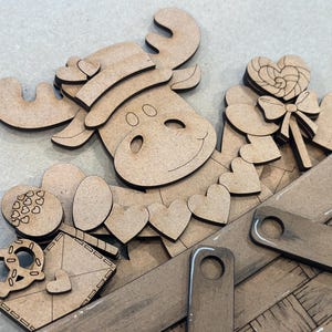 May include: A wooden craft kit featuring a moose with a hat, hearts, a candy cane, and a pretzel. The pieces are arranged in a wooden basket.