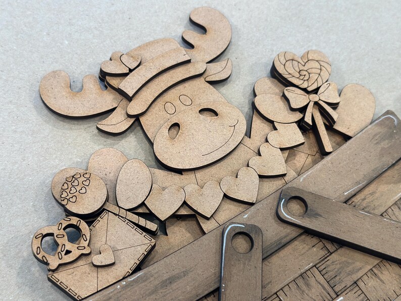 May include: A wooden craft kit featuring a moose with a hat, hearts, a candy cane, and a pretzel. The pieces are arranged in a wooden basket.