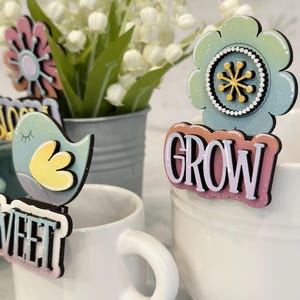 May include: A set of four colorful wooden plant markers with glitter accents. The markers feature a flower, a bird, and the words "Sweet" and "Grow".