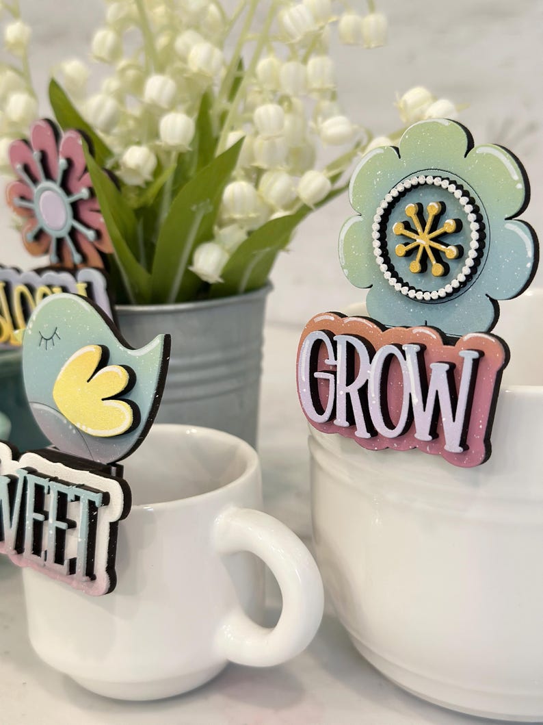 May include: A set of four colorful wooden plant markers with glitter accents. The markers feature a flower, a bird, and the words "Sweet" and "Grow".