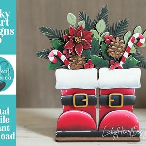 May include: A decorative wooden cutout featuring two red Santa boots with black belts and gold buckles, topped with a festive arrangement of a red poinsettia, pine branches, pinecones, red berries, and two candy canes, with Lucky Heart Designs Co text.