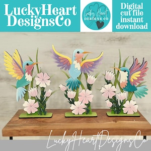 May include: Three colourful hummingbird cutouts with open wings, flowers, and greenery. The hummingbirds are painted in shades of blue, yellow, pink, and purple. The base is green, and the text "Lucky Heart Designs Co" is visible.