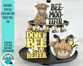 Puede incluir: Una bandeja de tres niveles decorada con un tema de vaca de las tierras altas y abejas. La bandeja presenta un letrero que dice "Don't Bee a Heifer", un letrero que dice "Bee-Moo-tiful", y un letrero con una vaca de las tierras altas y una abeja. La bandeja está decorada con pequeños recortes de abejas.