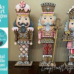 Può includere: Tre schiaccianoci natalizi in legno, ognuno con un design unico. Gli schiaccianoci sono dipinti in tonalità di marrone, rosa e bianco, con dettagli ispirati al pan di zenzero. L'immagine include il testo "Lucky Heart Designs Co" e "Digital cut file instant download."