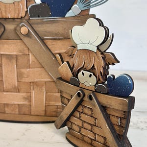 May include: A wooden figurine of a Highland cow wearing a chef's hat, holding a whisk and a spatula, standing in a brown wooden basket. The cow is brown and white with a fluffy mane. The basket has a brown and white checkered pattern.