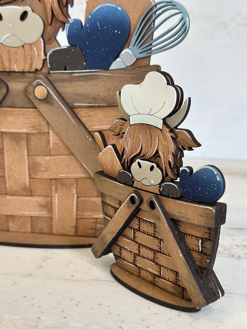 May include: A wooden figurine of a Highland cow wearing a chef's hat, holding a whisk and a spatula, standing in a brown wooden basket. The cow is brown and white with a fluffy mane. The basket has a brown and white checkered pattern.