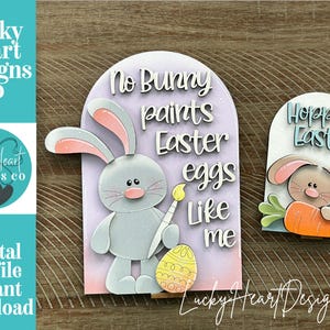 May include: Easter-themed wooden cutouts. One features a gray bunny painting an Easter egg, with the text "No Bunny paints Easter eggs like me." The other says "Hoppy Easter" with a bunny and carrot. The design includes the text "Lucky Heart Designs Co."