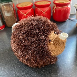 May include: A novelty tea cosy in the shape of a hedgehog. The cosy is made from brown, textured material, with a knitted, lighter brown snout and a white spout. The eyes are small and black. It sits on a dark grey countertop.