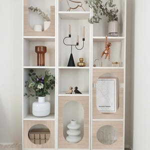 May include: A white and wood-effect display cabinet filled with decorative objects. The shelves feature vases, candles, a wooden monkey, and a map. The cabinet has a contemporary design with arched cutouts and is against a neutral wall.