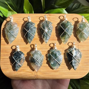 May include: Nine moss agate pendants with silver wire wrapping, displayed on a wooden tray. The pendants have a mix of green and clear tones, each with a unique pattern. The wire is wrapped around the stone, creating a cage-like design.
