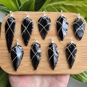 Op de afbeelding: Een verzameling van tien zwarte obsidiaan pijlpunten, elk omwikkeld met zilverkleurig draad. De hangers worden gepresenteerd op een lichtbruin houten bord, met groene bladeren op de achtergrond. Elke hanger is ongeveer 5 cm lang.
