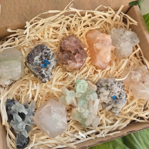 May include: A collection of various colorful crystals and minerals, including clear, pink, and green specimens, nestled in a cardboard box filled with straw-like packing material. The crystals vary in shape and size, showcasing natural textures and hues.