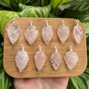 Può includere: Nove ciondoli in quarzo rosa pallido a forma di punta di freccia, ciascuno avvolto in filo color argento con un piccolo occhiello sulla parte superiore per appenderli. Le pietre hanno una consistenza cristallina traslucida su una tavola di legno.