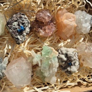 May include: A collection of various colorful crystals and minerals, including shades of pink, green, white, and gray. The crystals are arranged in a wooden box filled with straw-like packing material, creating a natural and earthy aesthetic.