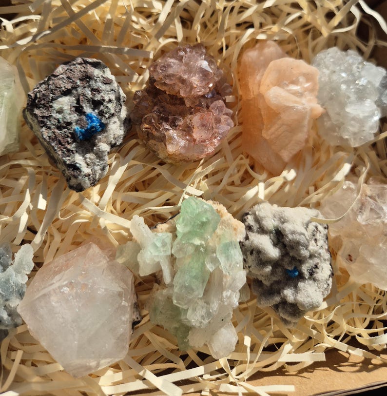 May include: A collection of various colorful crystals and minerals, including shades of pink, green, white, and gray. The crystals are arranged in a wooden box filled with straw-like packing material, creating a natural and earthy aesthetic.
