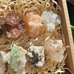 May include: A collection of raw crystals in a cardboard box filled with straw. The crystals vary in color, including shades of pink, peach, white, and green. The crystals have a rough, natural texture and are arranged in a visually appealing manner.