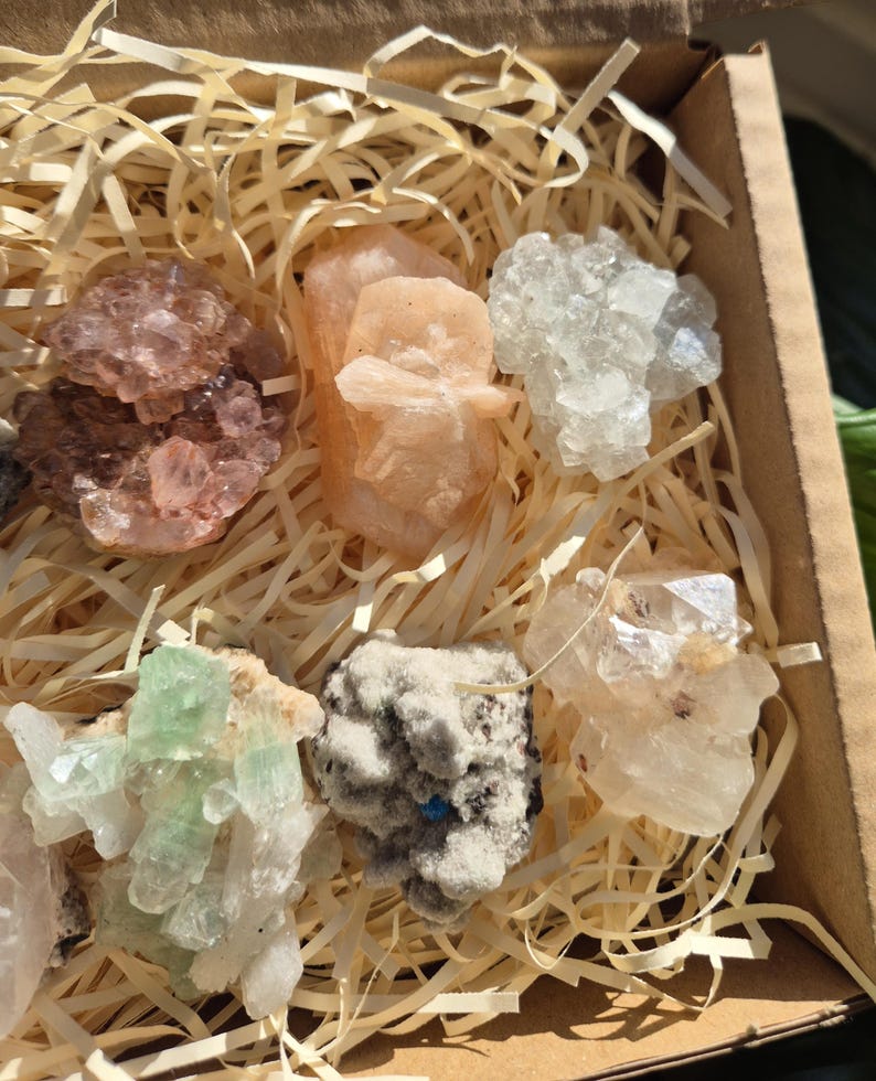 May include: A collection of raw crystals in a cardboard box filled with straw. The crystals vary in color, including shades of pink, peach, white, and green. The crystals have a rough, natural texture and are arranged in a visually appealing manner.