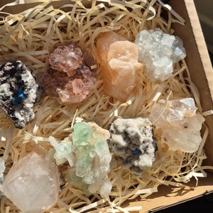 May include: A collection of various crystals and minerals in a cardboard box filled with straw-like packing material. The crystals display a range of colors, including pink, white, green, and gray. The box is open, revealing the assortment of natural gemstones.