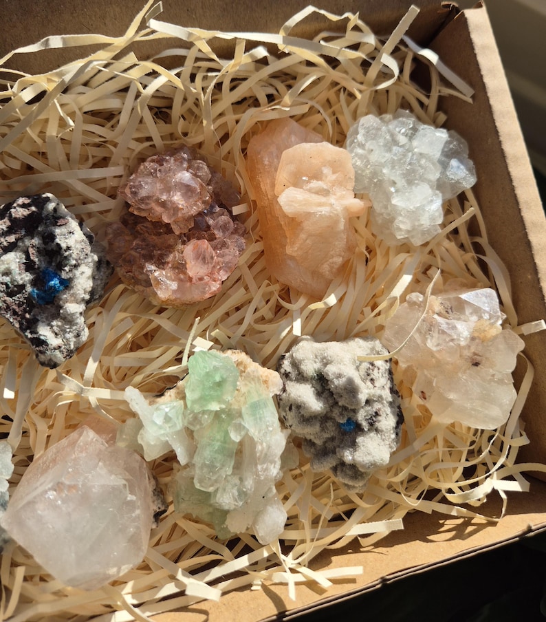 May include: A collection of various crystals and minerals in a cardboard box filled with straw-like packing material. The crystals display a range of colors, including pink, white, green, and gray. The box is open, revealing the assortment of natural gemstones.