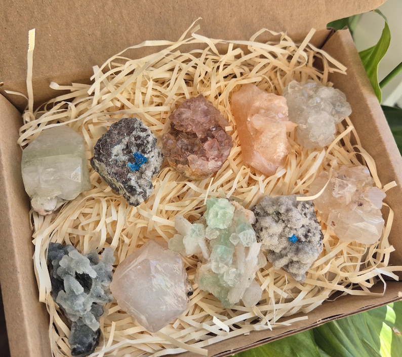 May include: A collection of various colorful crystals and minerals, including clear, pink, and green specimens, nestled in a cardboard box filled with straw-like packing material. The crystals vary in shape and size, showcasing natural textures and hues.