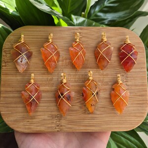 May include: Nine orange-red gemstone pendants, arrowhead-shaped and wrapped in gold wire, are arranged on a wooden board. The pendants have varying colours and textures. Green foliage forms the background.