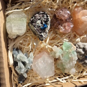 May include: A collection of various colorful crystals and minerals, including green, pink, and clear varieties, nestled in a cardboard box with straw-like packing material. The crystals exhibit diverse shapes and textures, showcasing natural geological formations.