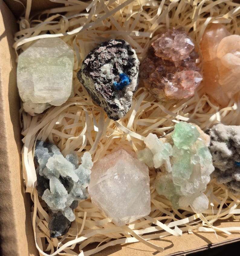 May include: A collection of various colorful crystals and minerals, including green, pink, and clear varieties, nestled in a cardboard box with straw-like packing material. The crystals exhibit diverse shapes and textures, showcasing natural geological formations.
