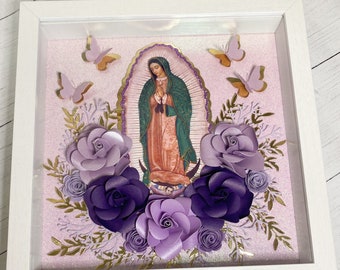 May include: A framed image of the Virgin Mary with a gold halo and a blue and green robe. The image is surrounded by paper flowers in shades of purple and lavender, and gold glitter.