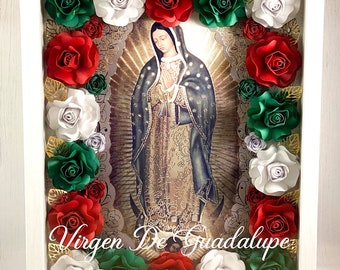 May include: A white frame with a picture of the Virgin of Guadalupe surrounded by paper flowers in red, white, and green. The text "Virgen De Guadalupe Ruega por nosotros" is written below the image.