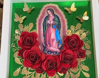 May include: A framed image of the Virgin Mary with a green background and red paper flowers surrounding the image. The flowers are arranged in a circle around the image and are accented with gold leaves.