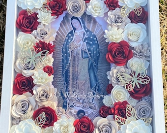 May include: A framed image of Our Lady of Guadalupe surrounded by paper flowers in shades of red, white, and beige. The flowers are arranged in a circular pattern around the image, creating a beautiful and intricate design.