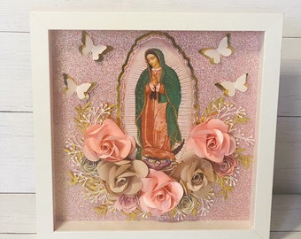 May include: A white framed shadow box with a pink glitter background. The box features a religious image of the Virgin Mary, surrounded by paper flowers in shades of pink and brown. White butterflies are also included in the design.