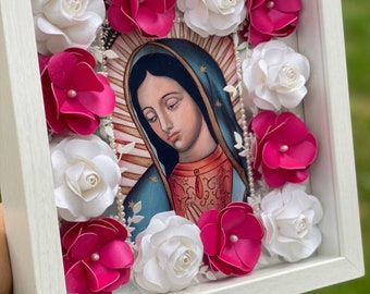 May include: A framed religious artwork featuring an image of the Virgin Mary, surrounded by white and fuchsia paper flowers. The artwork is set in a white frame, creating a three-dimensional effect. Decorative pearls and floral accents enhance the design.