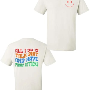 May include: Cream-coloured t-shirt featuring a red smiley face on the front and the text "ALL I DO IS TALK SHIT AND HAVE PANIC ATTACKS" in various colours on the back. A casual, unisex garment.
