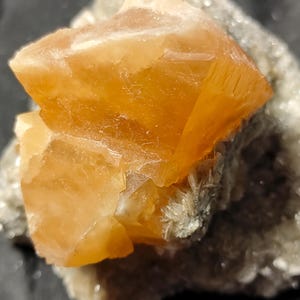 Könnte beinhalten: Nahaufnahme einer Mineralprobe mit einer großen, orange-gelben Kristallformation. Der Kristall hat ein geschichtetes Aussehen und sitzt auf einer Matrix aus kleineren, grau-weißen Kristallen. Die Beleuchtung hebt die Textur und Farbe des Kristalls hervor.