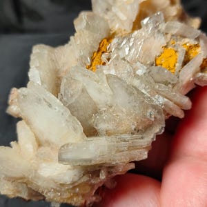 Puede incluir: Primer plano de un grupo de formaciones cristalinas translúcidas. Los cristales son principalmente blancos con toques de amarillo y naranja. El espécimen mineral tiene una apariencia rugosa y texturizada.