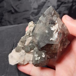 Könnte beinhalten: Ein Cluster aus rohen, natürlichen Kristallen in Grau-, Braun- und Weißtönen. Die Kristalle haben eine raue, strukturierte Oberfläche. Die Kristalle sind eine Mischung aus undurchsichtigen und durchscheinenden Abschnitten, wobei einige Bereiche dunkler erscheinen. Gehalten in einer Hand.