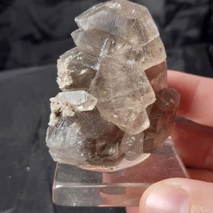 Könnte beinhalten: Ein Cluster aus Rauchquarzkristallen, die transluzente Brauntöne aufweisen. Die Kristalle sind auf einem transparenten, rechteckigen Sockel montiert. Die Quarzformation hat eine geschichtete Struktur mit mehreren Kristallspitzen.