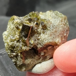 Op de afbeelding: Een close-up van een ruw, natuurlijk mineraal specimen. De steen heeft een mix van olijfgroene, bruine en roze tinten. Het oppervlak is getextureerd met kristallijne formaties en kleine, ingebedde kristallen. Het mineraal rust op een witte en roze ondergrond.
