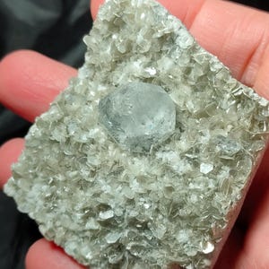 Op de afbeelding: Een lichtgrijs mineraal specimen met een centraal, lichtblauw kristal. De omringende matrix bestaat uit kleine, glinsterende, lichtgrijze vlokken. Het specimen wordt in een hand gehouden, wat de natuurlijke textuur en kleurvariaties benadrukt.