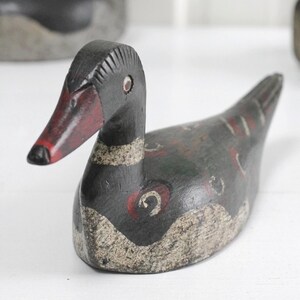 May include: A hand-carved wooden duck decoy featuring a dark black body with distressed white painted accents on the lower chest and wings. The duck has a red beak with a black tip, small circular eyes, and decorative red and white circular markings.