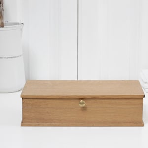 Puede incluir: Una caja rectangular de madera con un acabado liso de color marrón claro y un pequeño pomo redondo de latón centrado en el panel frontal. La caja presenta una tapa plana y un diseño sencillo, colocada horizontalmente sobre una superficie.