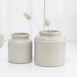 Op de afbeelding: Twee lichtgrijze keramische potten van verschillende formaten staan op een wit oppervlak. Eén pot heeft een kleine opening, de andere een bredere opening met gedroogde bloemen. Een gevouwen linnen doek en decoratieve objecten staan op de achtergrond.