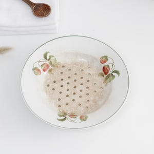 Può includere: Una ciotola scolabacche in ceramica bianca con un delicato motivo di fragole e foglie verdi lungo il bordo. La ciotola presenta una base circolare con numerosi fori di drenaggio distribuiti uniformemente per sciacquare frutta fresca.