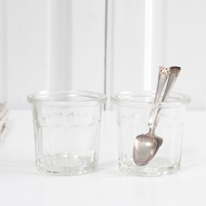 May include: Two clear glass jars with a textured design, one holding two silver spoons. A small white ceramic garlic and a decorative white ceramic bulb sit on a folded beige cloth.