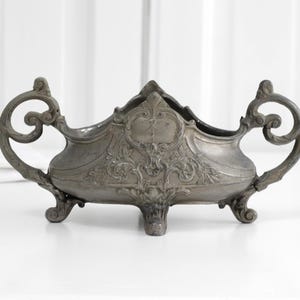 Op de afbeelding: Een metallic grijze sierschaal met gedetailleerd krulwerk en florale reliëfversieringen. De schaal heeft twee grote, gebogen handvatten aan weerszijden en rust op drie decoratieve pootjes, afgewerkt met een donkere, verweerde uitstraling.